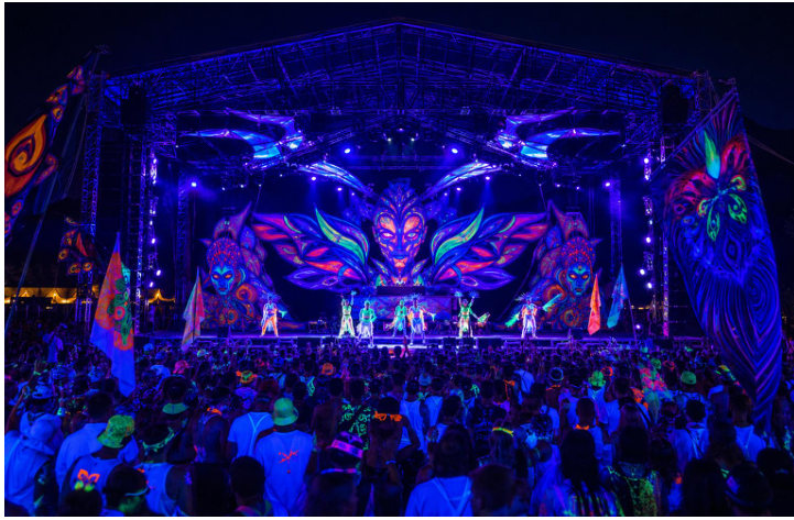 Outdoor night festival stage washed in UV, with fluorescent set pieces and a crowd